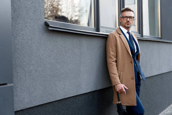 Bell'uomo d'affari in occhiali che tiene l'ombrello e in piedi in cappotto beige vicino all'edificio — Foto stock