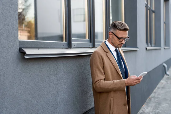 Homme d'affaires confiant dans des lunettes regardant smartphone tout en se tenant en manteau beige près du bâtiment — Photo de stock