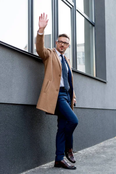 Fröhlicher Geschäftsmann mit Brille, winkende Hand im Stehen mit überkreuzten Beinen im beigen Mantel — Stockfoto