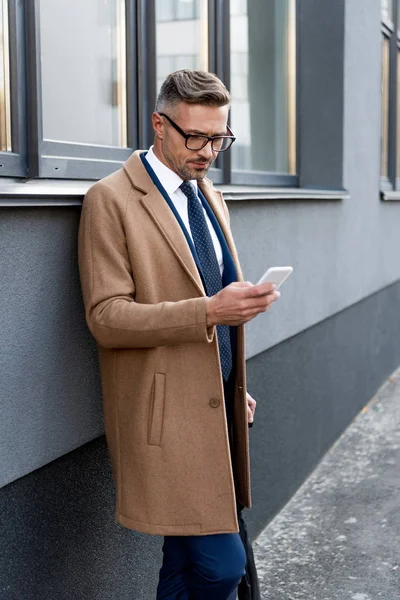 Homme d'affaires sérieux en lunettes regardant smartphone tout en se tenant en manteau beige près du bâtiment — Photo de stock