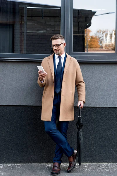 Alegre hombre de negocios en gafas mirando el teléfono inteligente mientras está de pie en abrigo beige y con paraguas - foto de stock