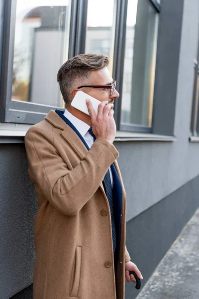 Handsome businessman in glasses talking on smartphone while standing in beige coat — Stock Photo