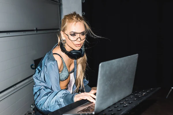 Atractiva mujer dj en gafas de pie y mirando el ordenador portátil - foto de stock