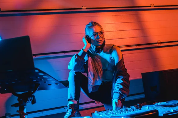 Thoughtful dj woman in glasses touching dj mixer while listening music in headphones — Stock Photo