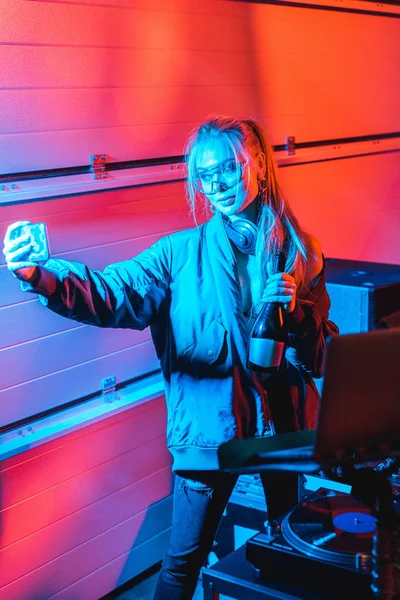 Elegante rubia dj mujer sosteniendo botella y tomando selfie en discoteca - foto de stock