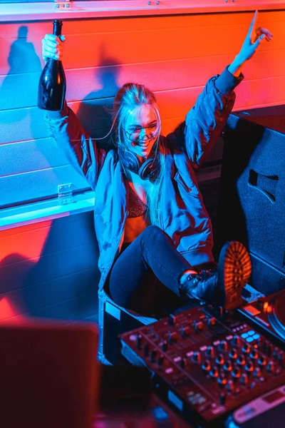 Alegre dj mujer en auriculares celebración de botella y gesto mientras está sentado en discoteca - foto de stock