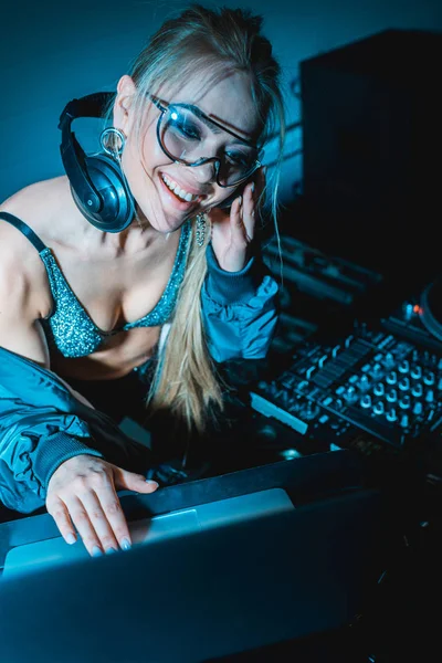 Cheerful blonde dj girl listening music in headphones and using laptop in nightclub — Stock Photo