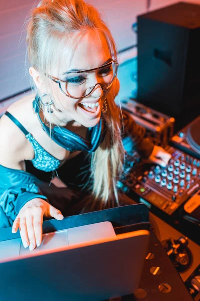 Cheerful blonde dj girl in headphones using laptop in nightclub — Stock Photo