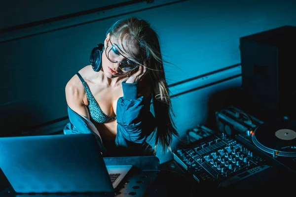 Beautiful blonde dj girl listening music in headphones and using laptop in nightclub — Stock Photo