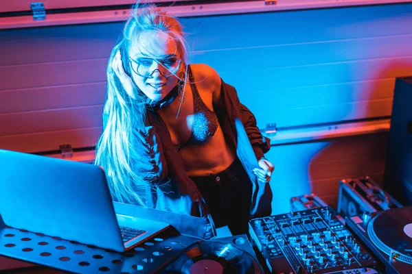 Beautiful and cheerful dj woman listening music in headphones and using laptop in nightclub — Stock Photo