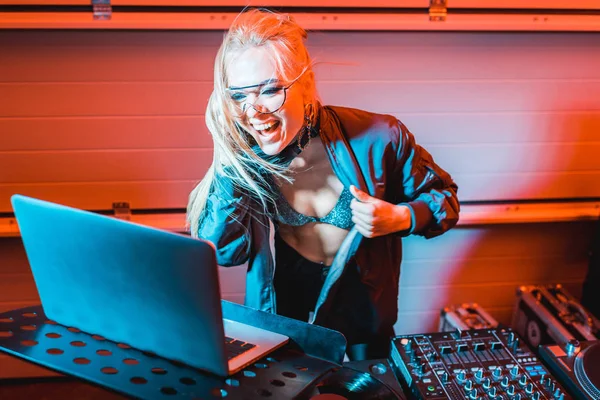 Schöne und glückliche DJ-Frau, die Musik über Kopfhörer hört und Laptop im Nachtclub benutzt — Stockfoto