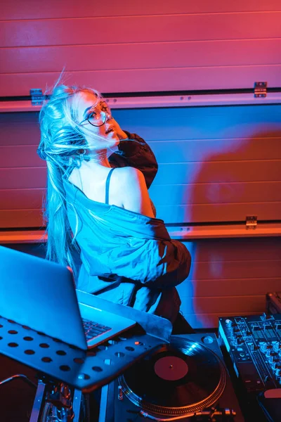 Fröhliche DJ-Frau mit Kopfhörern, die in der Nähe von Laptop in Nachtclub steht — Stockfoto