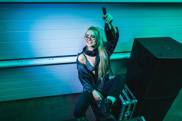 Cheerful dj girl holding bottle with wine in hand and smiling in nightclub — Stock Photo