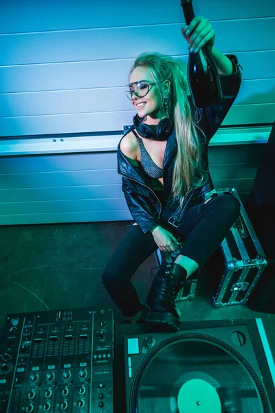 Cheerful dj girl in glasses holding bottle with wine in hand and smiling in nightclub — Stock Photo