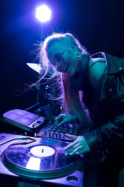 Feliz rubia dj chica tocando vinilo registro y sonriendo en nightclub - foto de stock