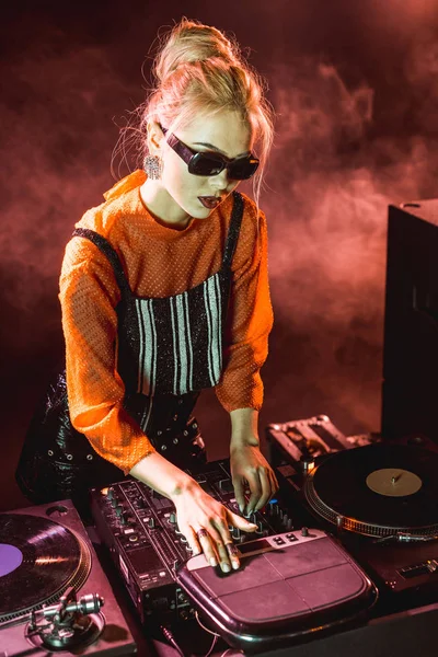 Blonde dj fille en lunettes de soleil touchant équipement dj dans la boîte de nuit avec de la fumée — Photo de stock