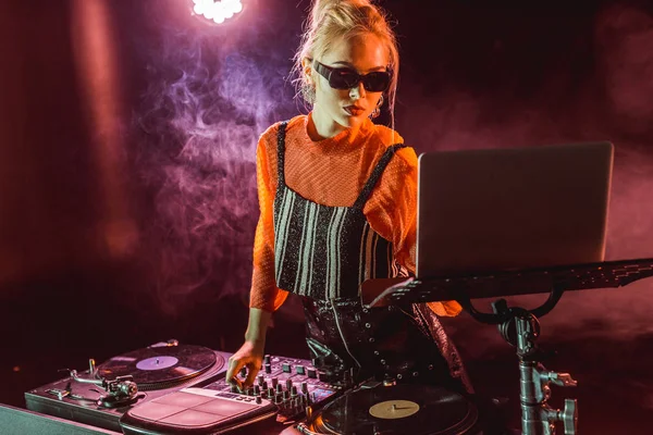Elegante chica dj en gafas de sol tocando equipo dj mientras mira el ordenador portátil en discoteca con humo - foto de stock
