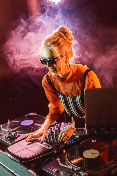 Весела дівчина джи в сонцезахисних окулярах, що торкаються обладнання джи в нічному клубі з димом — стокове фото