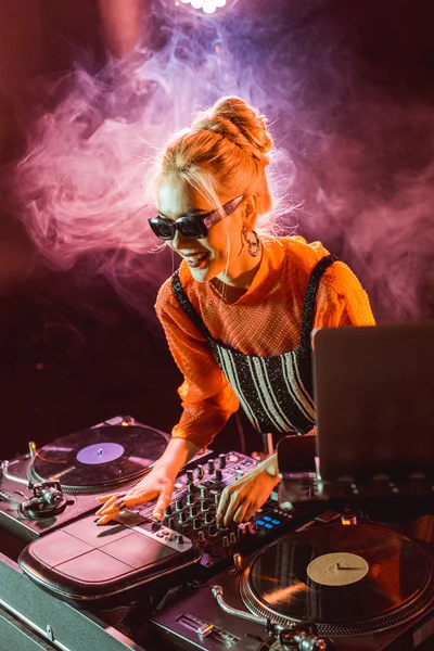 Alegre dj menina em óculos de sol tocando equipamento dj em boate com fumaça — Fotografia de Stock