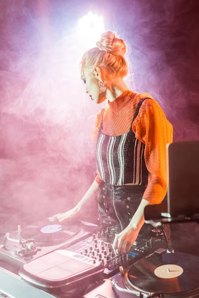 Blonde dj girl touching dj equipment while standing near laptop in nightclub with smoke — Stock Photo
