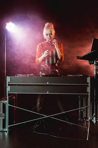 Atractiva y elegante chica dj en los auriculares mirando dj mezclador en discoteca con humo — Stock Photo