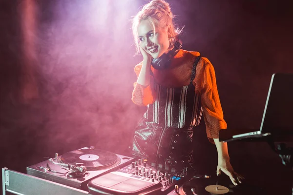 Happy and stylish dj girl in headphones standing near dj mixer in nightclub with smoke — Stock Photo