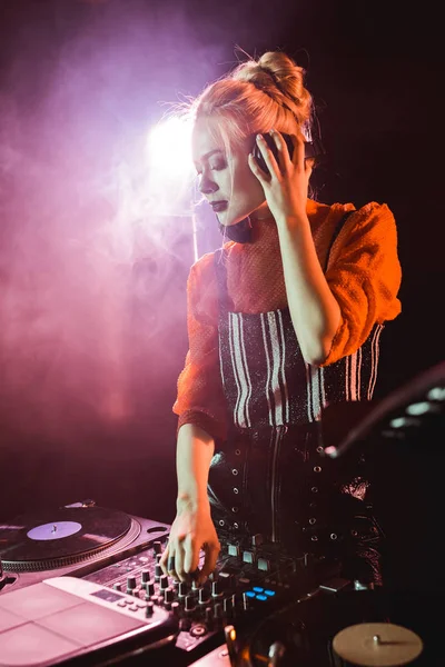 Stylish dj girl in headphones standing near dj mixer in nightclub with smoke — Stock Photo