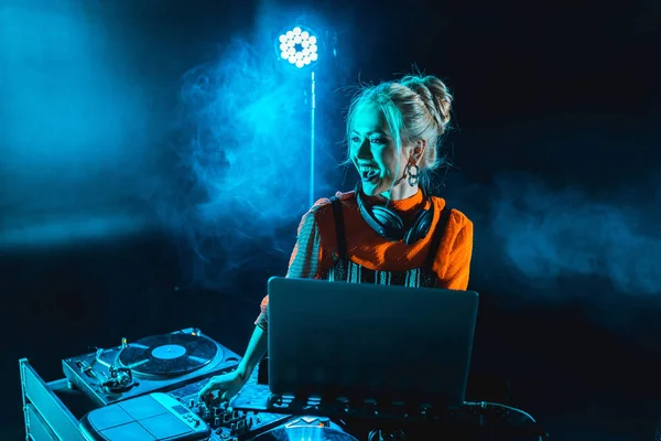 Happy dj girl in headphones standing near dj mixer and laptop in nightclub with smoke — Stock Photo