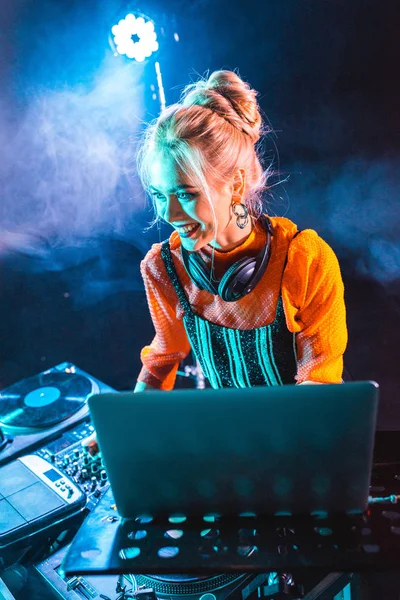 Sonriente chica dj en los auriculares de pie cerca de dj mezclador y portátil en discoteca con humo - foto de stock