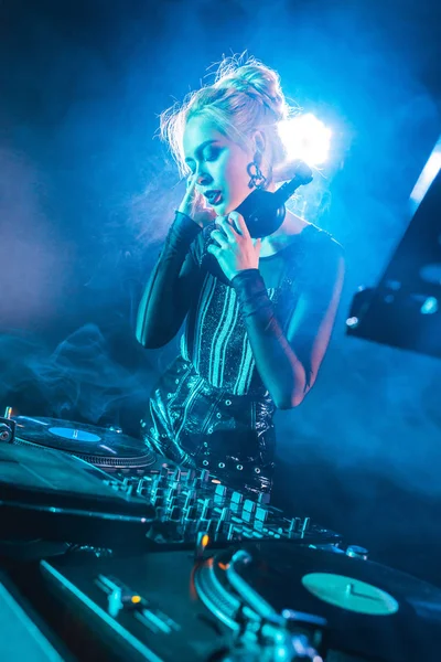 Attractive dj girl standing near dj mixer and touching headphones in nightclub with smoke — Stock Photo