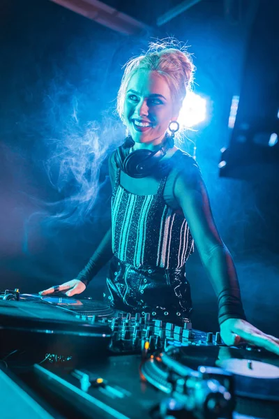 Happy dj girl standing near dj mixer and touching vinyl records in nightclub with smoke — Stock Photo