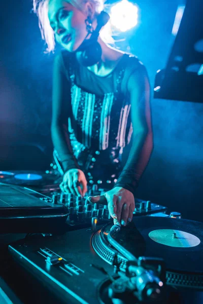 Selective focus of blonde dj girl standing near dj mixer and touching vinyl records in nightclub with smoke — Stock Photo