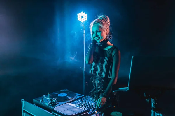 Happy blonde dj girl in headphones standing near dj mixer in nightclub with smoke — Stock Photo