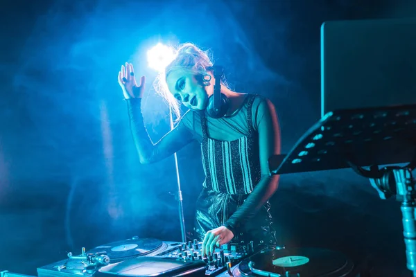 Smiling blonde dj girl in headphones standing near dj mixer in nightclub with smoke — Stock Photo