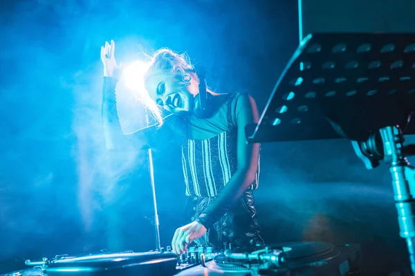 Enfoque selectivo de la chica feliz dj en los auriculares de pie cerca de dj mezclador en discoteca con humo — Stock Photo