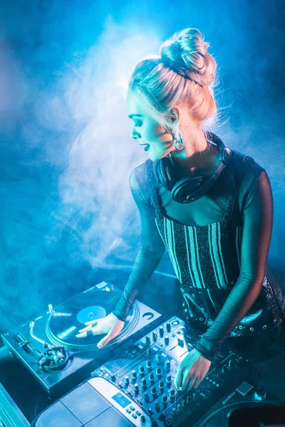 Счастливая блондинка в наушниках, стоящая рядом с ди-джеем и виниловой пластинкой в ночном клубе с дымом — стоковое фото