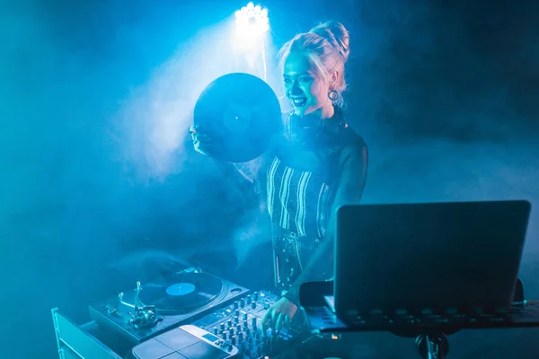 Glückliches blondes dj Mädchen, das neben dj Ausrüstung steht und Retro-Schallplatte in Nachtklub mit Rauch hält — Stockfoto