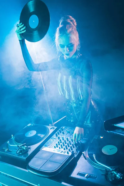 Blonde dj girl looking at dj equipment and holding retro vinyl record in nightclub with smoke — Stock Photo