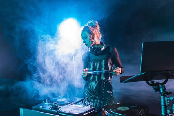 Atraente dj mulher olhando para equipamento dj e segurando recorde de vinil retro em boate com fumaça — Fotografia de Stock