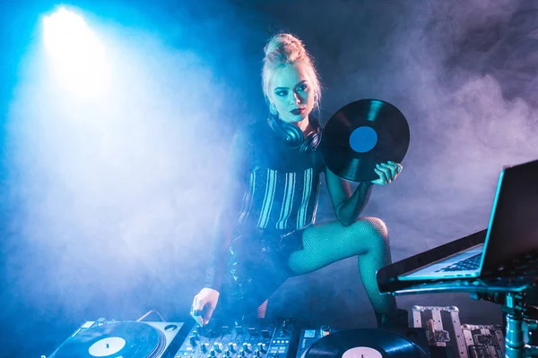 Attractive dj woman holding retro vinyl record near dj equipment in nightclub with smoke — Stock Photo