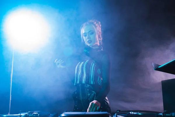 Blonde dj woman holding retro vinyl record and looking at dj equipment in nightclub with smoke — Stock Photo