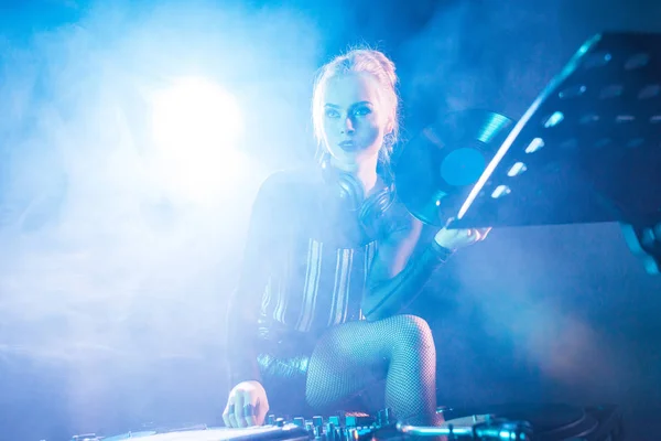 Elegante dj mulher segurando recorde de vinil retro e de pé perto do equipamento dj em boate com fumaça — Fotografia de Stock