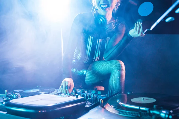 Abgeschnittene Ansicht einer fröhlichen DJ-Frau, die eine Retro-Schallplatte in der Hand hält und in der Nähe von DJ-Geräten in einem Nachtclub mit Rauch steht — Stockfoto