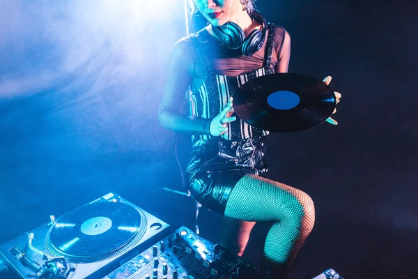 Vista recortada de la mujer dj sosteniendo registro de vinilo retro y de pie cerca de equipo dj en discoteca con humo - foto de stock