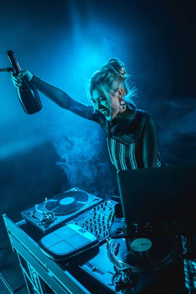 Cheerful blonde dj woman holding  bottle and smiling near dj equipment in nightclub with smoke — Stock Photo