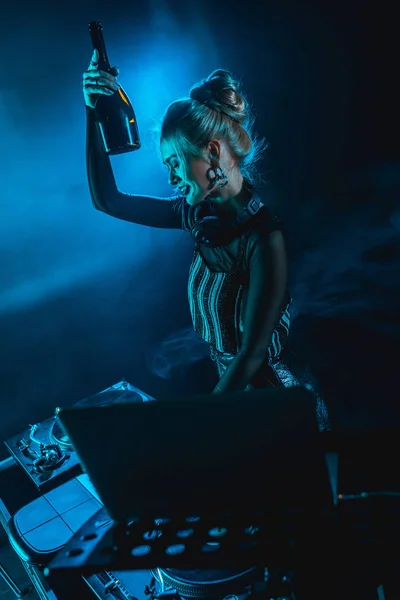 Fröhliche blonde dj Frau mit Kopfhörer, Flasche in der Hand und lächelnd in der Nähe dj Ausrüstung in Nachtclub mit Rauch — Stockfoto