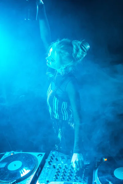 Счастливая блондинка-ди-джей в наушниках держит бутылку рядом с диджейским оборудованием в ночном клубе с дымом — стоковое фото