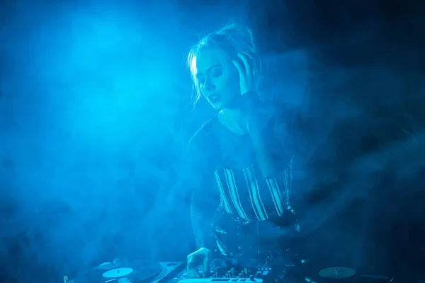 Blonde dj girl listening music in headphones near dj equipment in nightclub with smoke — Stock Photo