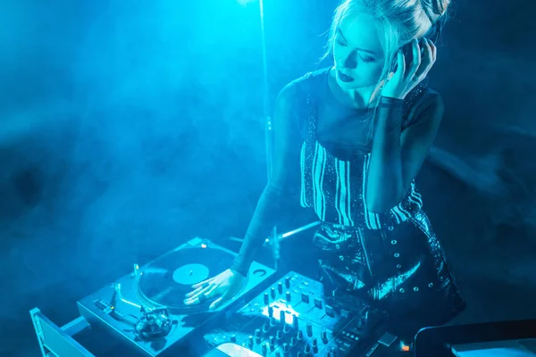 Blonde dj girl listening music in headphones while looking at dj equipment in nightclub with smoke — Stock Photo
