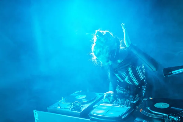 Blonde dj girl listening music in headphones while using dj equipment in nightclub with smoke — Stock Photo